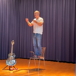 Oliver Steller steht auf einem Stuhl auf der Bühne in der Aula. Neben ihm steht eine silberfarbene Gitarre.