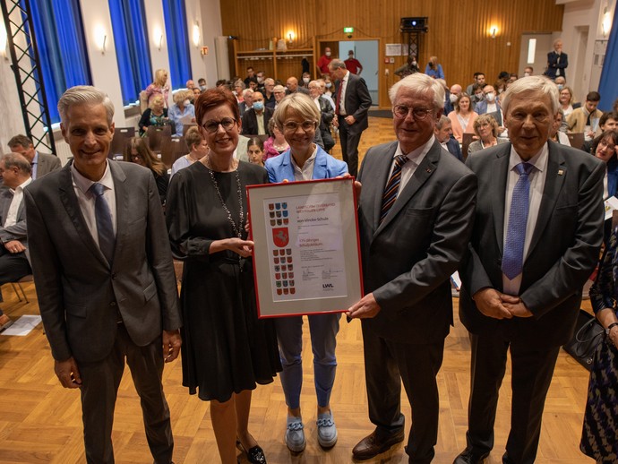 Vorne: Andreas Liebald, Birgit Westers, Dorothee Feller, Klaus Baumann (mit Urkunde), Dr. Günter Fiedler. Hinten: Gäste des Festakts in der Aula (vergrößerte Bildansicht wird geöffnet)