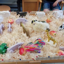 Ein Stall mit Holzwolle ausgelegt, darin zahlreiche Holztiere: Katzen, Schweine, Papagei, Giraffe, Ziege, Pferd, Frosch... (vergrößerte Bildansicht wird geöffnet)