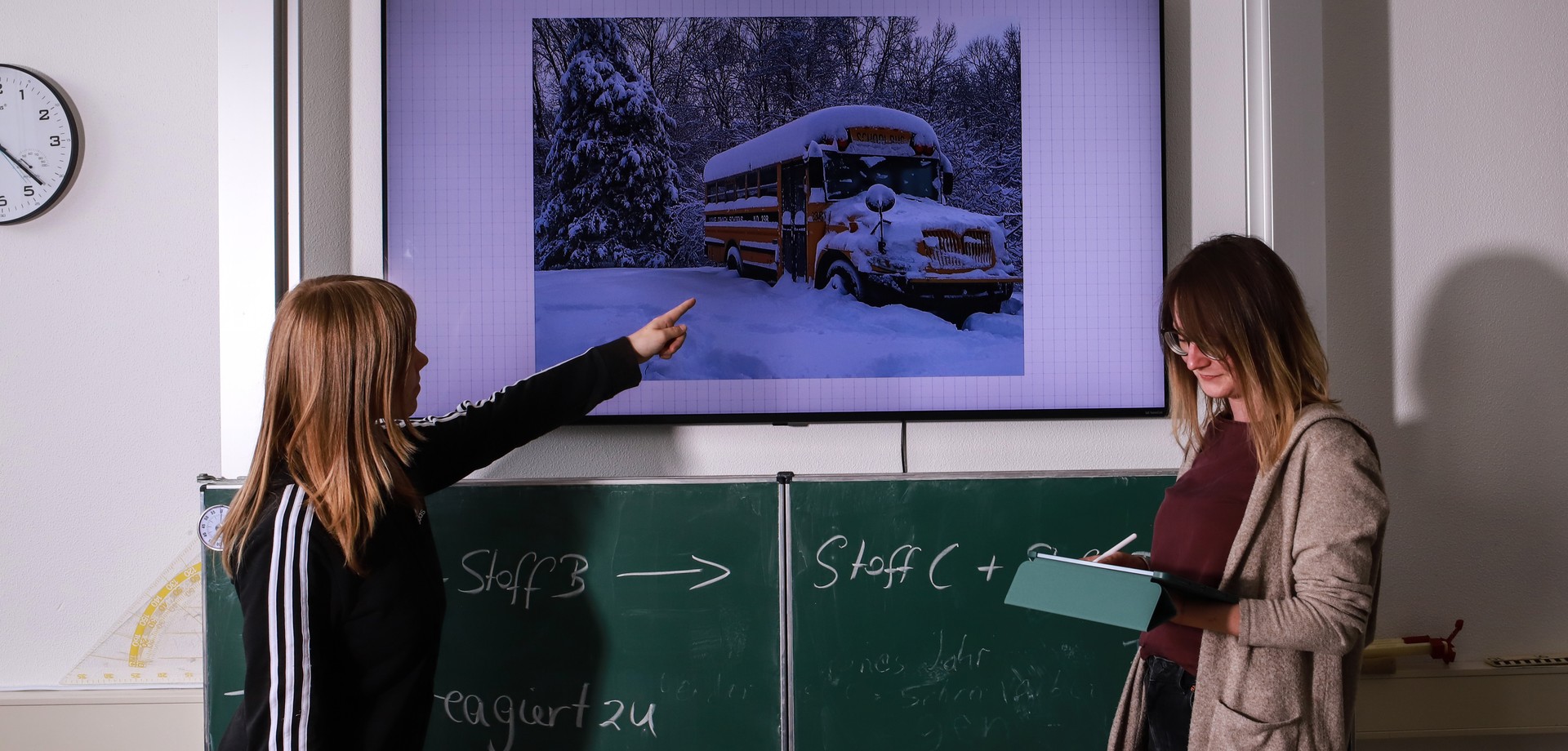 Zwei Schülerinnen stehen vor einer Tafel, eine zeigt auf ein Bild über der Tafel