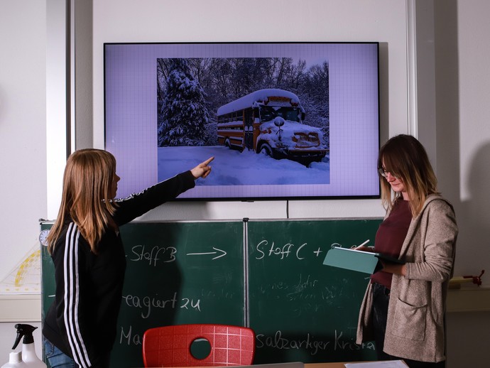 Zwei Schülerinnen stehen vor der Tafel, eine zeigt auf ein Bild über der Tafel