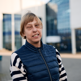 Portrait Sabrina Führer, blonde kurze Haare, blaue Weste, blau-weißer Pullover, im Hintergrund die Glasfassade des Uni-Gebäudes